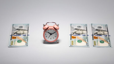 A pink alarm clock sits between two stacks of hundred dollar bills. The clock shows time, emphasizing the relationship between time and money in a minimalist concept.の写真素材