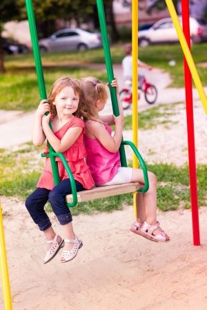 Two beautiful little girls on a swings outdoor in the playground at summertimeの写真素材