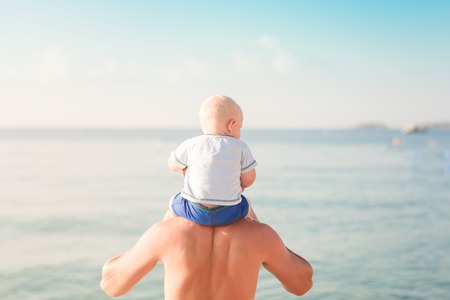 Happy baby sitting on the father's shoulders at the beach looking at the seaの写真素材