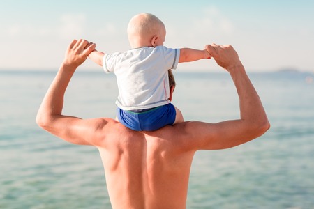 Happy baby sitting on the father's shoulders at the beach looking at the seaの写真素材
