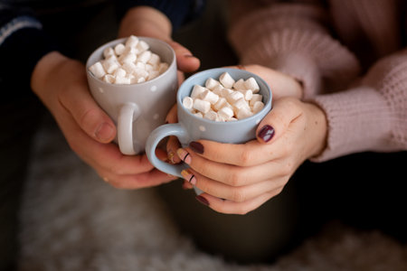 Happy couple's hands holding hot cups with hot drinks with marshmallow. Comfort and coziness of home...