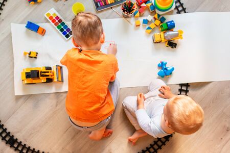 Preschool boys drawing on floor on paper, playing with educational toys - blocks, train, railroad, vehicles at home or daycare. Toys for preschool and kindergarten. Top view. Children's art and creativeness conceptの写真素材
