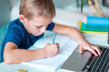 Closeup of little young school boy working at home with a laptop and class notes studying in a...