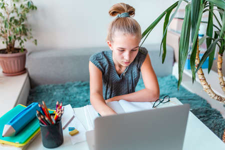 Beautiful young school girl left-handed working at home in her room with laptop and class notes studying in a virtual class. Distance and online education, e-learning during quarantineの写真素材
