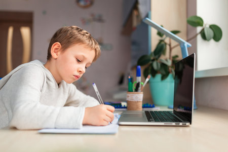 Cute little schoolboy studying at home doing school homework. Training books and notebook on the table. Distance learning online educationの写真素材