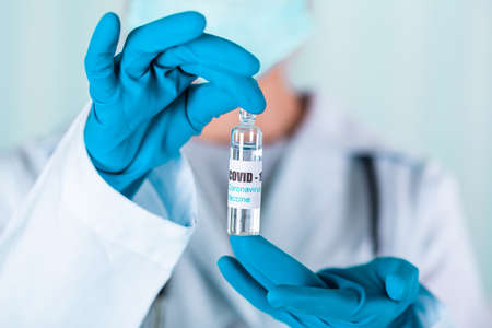 Woman doctor or nurse in uniform and gloves wearing face mask protective in lab, holding medicine vial vaccine bottle with COVID-19 Coronavirus vaccine labelの写真素材