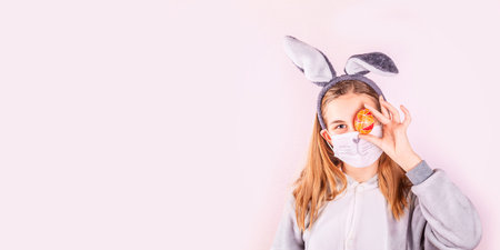 Girl in rabbit bunny ears on head and protective mask with colored eggs on pink background. Cheerful smiling happy child.  Easter holiday bannerの写真素材