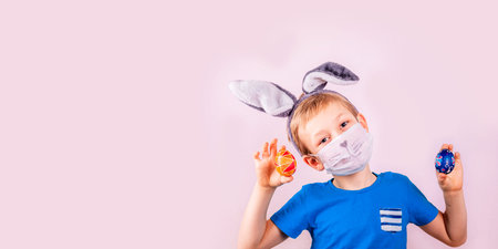 Cute little boy in rabbit bunny ears on head and protective mask with colored eggs on pink...