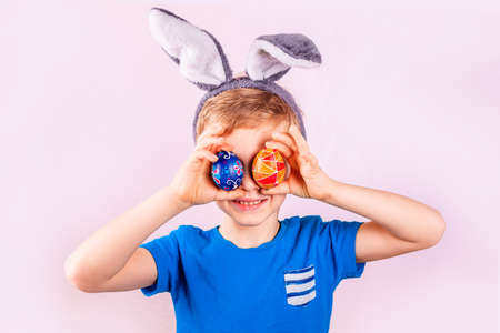 Cute little boy in rabbit bunny ears on head closing his eyes with colored eggs on pink background. Cheerful smiling happy child. Easter holidayの写真素材