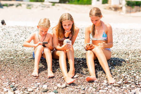 Children playing games and searching web on the telephones at the beach. Gadget dependency disorder problem for kids during holiday vacation at the seaside conceptの写真素材