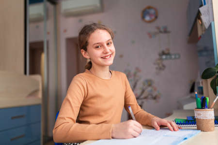 Beautiful smiling schoolgirl studying at home doing school homework. Training books and notebook on the table. Distance learning online educationの写真素材
