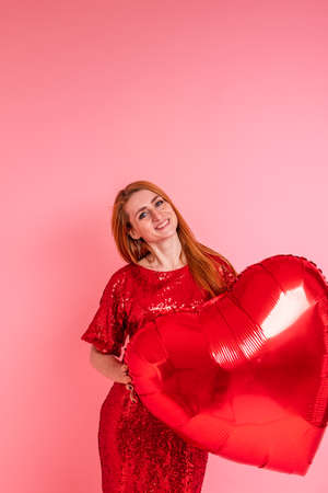 Beautiful redhead girl with red heart baloon posing. Happy Valentine's Day concept. Studio photo of beautiful ginger girl dancing on pink background.の写真素材