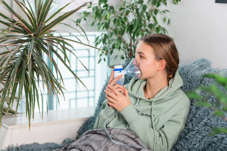 Teenage girl makes inhalation with a nebulizer equipment. Sick child holding inhalator in hand and breathes through an inhaler at home. Physical therapy for cold, flu and bronchial asthmaの写真素材