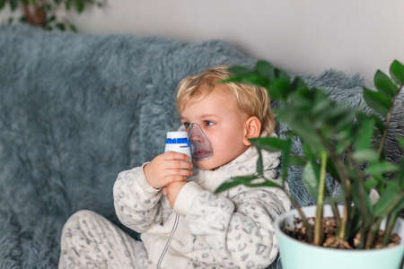 Cute baby boy makes inhalation with a nebulizer equipment. Sick child holding inhalator in hand and breathes through an inhaler at home. Physical therapy for cold, flu and bronchial asthmaの写真素材