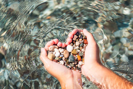 Hands full of colorful pebbles forming heart shape on the sea waves background. Holiday, rest, vacation at the seaside conceptの写真素材