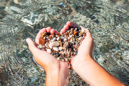 Hands full of colorful pebbles forming heart shape on the sea waves background. Holiday, rest, vacation at the seaside conceptの写真素材