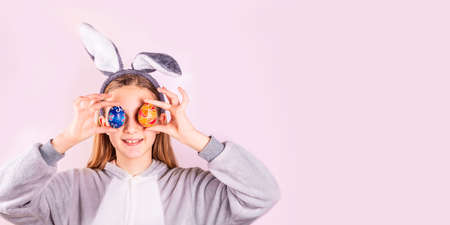 Girl in rabbit bunny ears on head with colored eggs on pink background. Cheerful smiling happy child. Easter holiday bannerの写真素材
