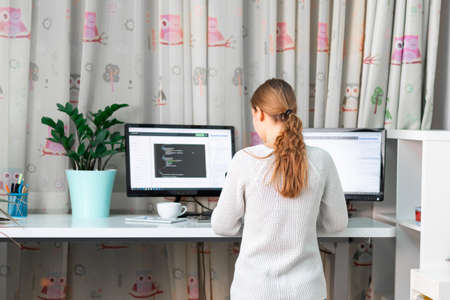 Young girl working on computer at standing desk at home office. Distance education, e-learning working from home. High standing desk table comfortable for healthy backの写真素材