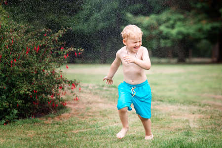 Little kid playing with water on hot summer day. Boy with garden sprinkler. Outdoor fun. Boy runs on a field under water drops.の写真素材