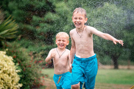 Kids play with water on hot summer day. Children with garden sprinkler having fun. Outdoor fun. Boys run on a field under water drops.の写真素材