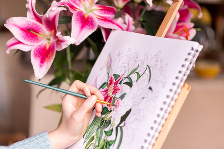 Young left-handed girl painting still life with flowers, purple lilies, with watercolor paints on the easel at homeの写真素材