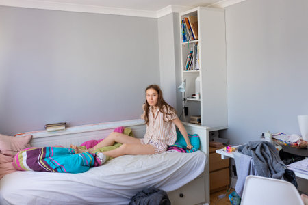 A teenage girl in pajamas sits in bed, looking shocked at the mess in her cluttered bedroom....