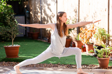 A teenage girl performing Virabhadrasana, the warrior pose, on a yoga mat in her backyard. Outdoor yoga practice showing strength, balance, and a healthy lifestyle.の写真素材