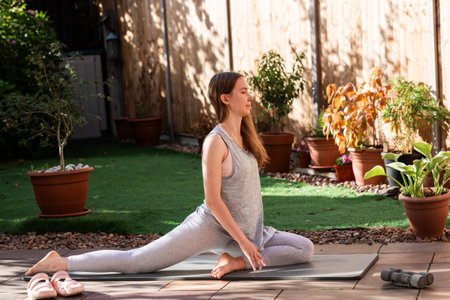 A teenage girl practicing Pigeon Pose (Eka Pada Rajakapotasana) on a yoga mat in her backyard. Outdoor yoga session focusing on hip flexibility, balance, and a healthy, mindful lifestyle.の写真素材