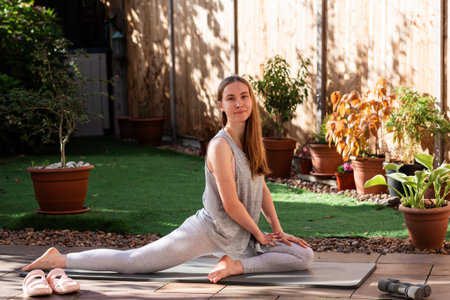 A teenage girl practicing Pigeon Pose (Eka Pada Rajakapotasana) on a yoga mat in her backyard. Outdoor yoga session focusing on hip flexibility, balance, and a healthy, mindful lifestyle.の写真素材
