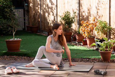A teenage girl performing a seated spinal twist on a yoga mat in her backyard garden. Gentle yoga stretch promoting spinal mobility, flexibility, and a mindful outdoor practice.の写真素材