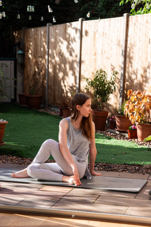 A teenage girl performing a seated spinal twist on a yoga mat in her backyard garden. Gentle yoga stretch promoting spinal mobility, flexibility, and a mindful outdoor practice.の写真素材