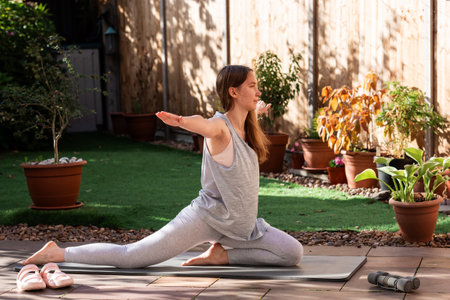A sporty teenage girl doing fitness and yoga exercises on a mat in her backyard garden. Outdoor activity with dumbbells, casual lifestyle, and focus on health.の写真素材