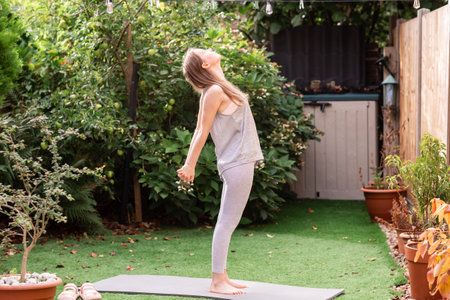 Teenage girl doing stretching exercises in a sunny backyard. Active young woman warming up before workout, enjoying outdoor fitness and fresh air. Concept of healthy lifestyle, mindfulness, flexibility, and wellbeing at home in a natural environment.の写真素材