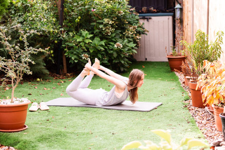 Teenage girl performing Dhanurasana (Bow Pose) on a yoga mat in the garden. Young woman stretching...