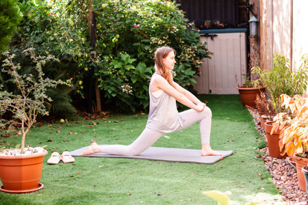 Teenage girl doing stretching exercises in a sunny backyard. Active young woman warming up before workout, enjoying outdoor fitness and fresh air. Concept of healthy lifestyle, mindfulness, flexibility, and wellbeing at home in a natural environment.の写真素材