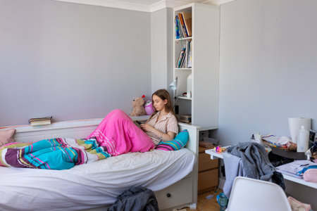 A teenage girl in pajamas lies in bed with her mobile phone in a messy room. Clothes, clutter, and an unmade bed reflect a typical teenager's everyday lifestyleの写真素材