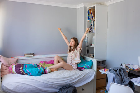 A teenage girl in pajamas lies in bed yawning and stretching. The cozy morning scene shows natural...