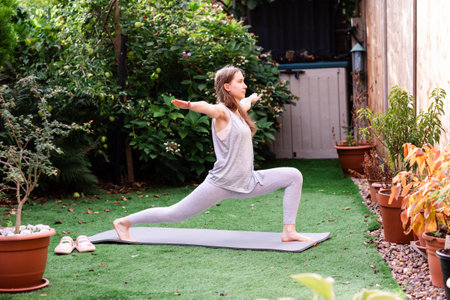 Teenage girl practicing Virabhadrasana (Warrior Pose) on a yoga mat in a sunny backyard. Confident young woman balancing and stretching during outdoor yoga session. Concept of mindfulness, focus, strength, and healthy lifestyle in a calm natural environment.の写真素材