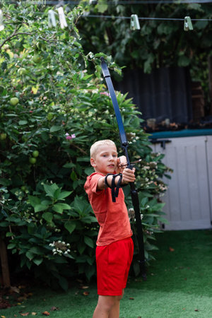 A young boy stands in a sunny back garden, practicing archery with a bow and arrow. Surrounded by...