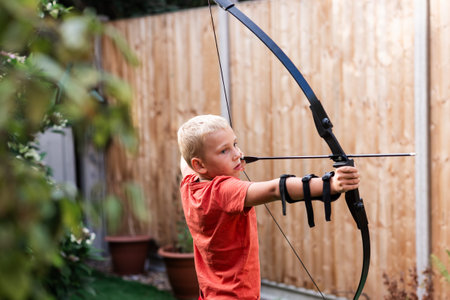 A young boy stands in a sunny back garden, practicing archery with a bow and arrow. Surrounded by...