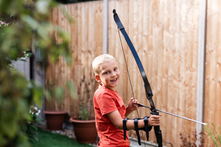 A young boy stands in a sunny back garden, practicing archery with a bow and arrow. Surrounded by...