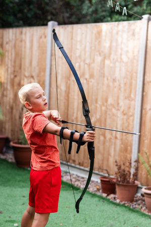 A young boy stands in a sunny back garden, practicing archery with a bow and arrow. Surrounded by green grass and flowers, he focuses carefully on his target. The scene captures concentration, skill-building, and the joy of outdoor play in a peaceful home setting.の写真素材