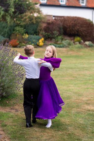 A young boy and girl gracefully perform a ballroom dance in a lovely garden. Dressed elegantly, they move with poise and joy under the sunlight, surrounded by flowers and greenery. The moment captures youthful elegance, teamwork, and the beauty of dance in a natural, outdoor settingの写真素材