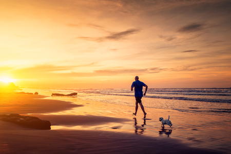 Adult man and small maltese dog are running at the beach in sunset timeの写真素材