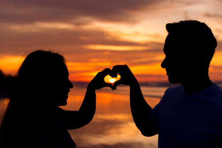 silhouette of couple doing heart shape by fingers against sunsetの写真素材