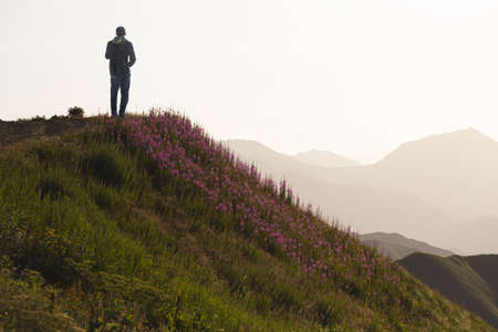A young man tourist with a backpack admires the sunset at dawn in the mountains against the background of flowering slopes. Distant mountain chains with a haze of sunlightの写真素材
