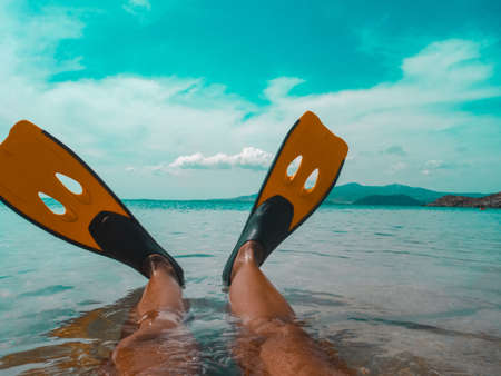 Women's legs in swimming fins in the aquamarine tropical sea. Hot sunny summer tourist season. Mountain range on the horizon, white clouds in the skyの写真素材