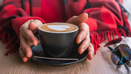 A woman wrapped in a red checkered blanket holds a hot cup of coffee in her hands. Cozy scene. Close-up of female middle-aged handsの写真素材