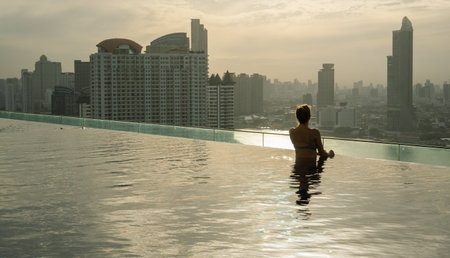 A woman sits in an infinity pool, gazing at the Bangkok skyline during sunset. The view showcases modern skyscrapers and soft golden light reflecting off the water, creating a serene and urban atmosphere.の写真素材