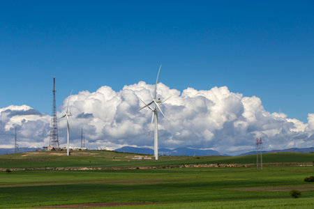 A scenic landscape featuring towering wind turbines amidst lush green fields under a bright blue sky. Mountains rise in the distance, framed by fluffy white clouds. A vision of clean energy and nature's harmonyの写真素材